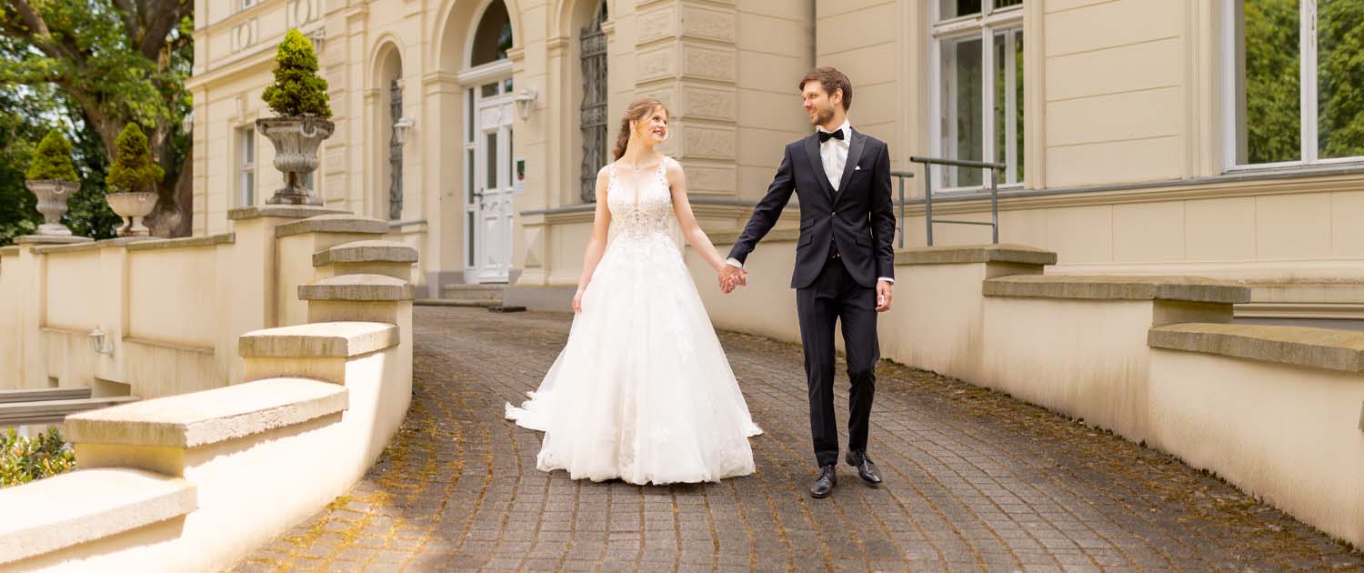 Anne und Julian gehen Hand in Hand die Auffahrt vor dem Eventschloss Schönfeld hinunter, schauen sich lächelnd an, umgeben von der historischen Kulisse des Schlosses.