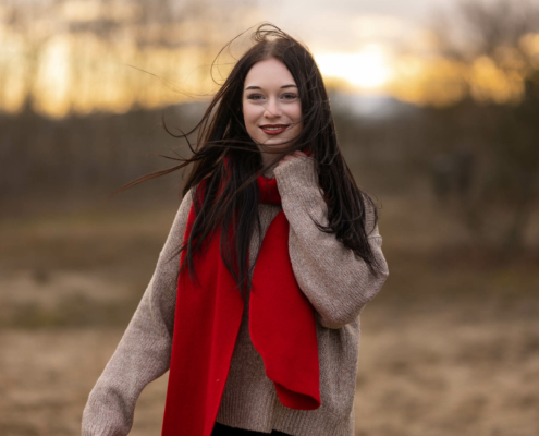 Mara Luisa hält ihren roten Schal am Hals fest, während der Wind ihre langen, dunklen Haare durch das Bild wirbelt, auf einem offenen Feld in Stendal.