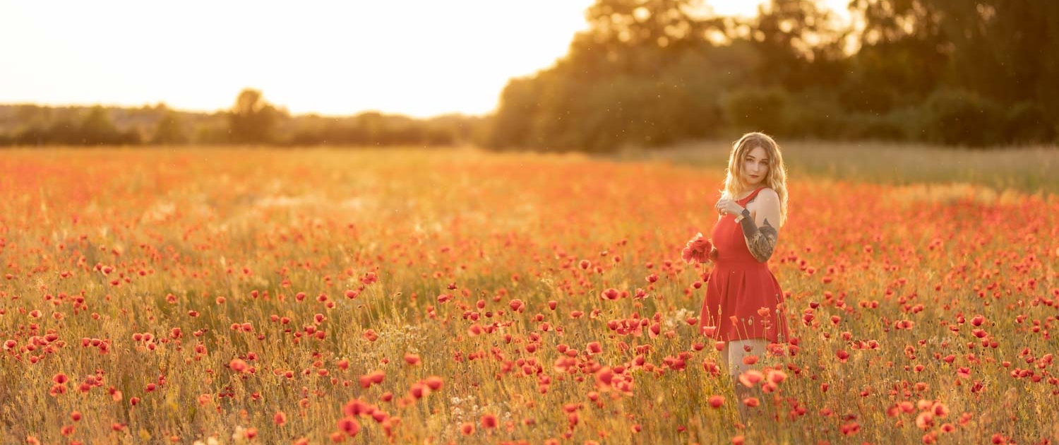 Michelle steht seitlich im Mohnfeld, hält Mohnblumen vor dem Bauch, blickt in die Kamera, umgeben von blühenden Mohnblumen, als Titelbild des Fotoshootings.