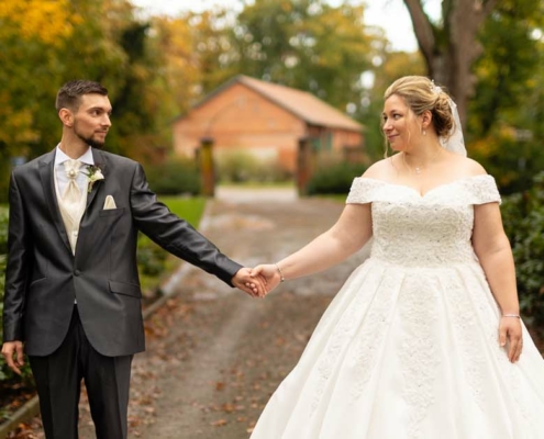 Gloria und Thomas stehen Hand in Hand auf einem Weg zum Schloss, umgeben von Hecken, schauen sich an, während ihrer Herbsthochzeit.